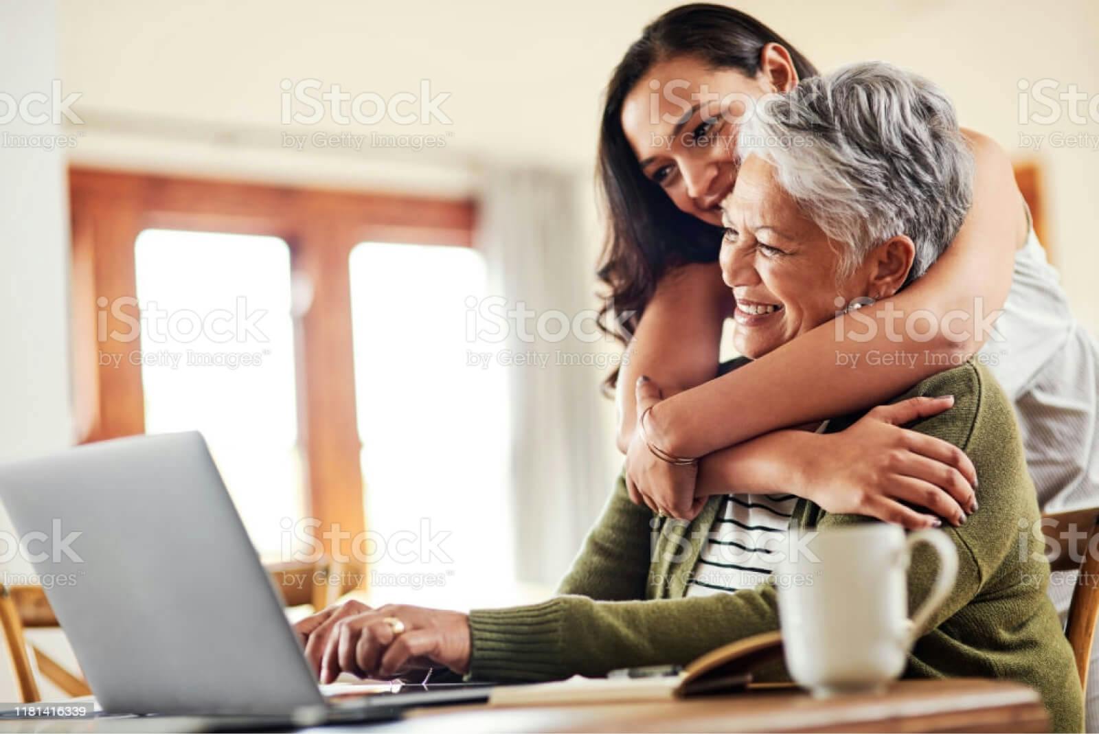 Watermarked stock image of a woman hugging her mother as they work on her laptop